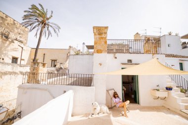 Güneşli bir çatıda bir kitapla rahatlayan bir kadın, kumaş bir gölgeyle gölgelenmiş, yanında büyük beyaz bir köpek, büyüleyici bir güney İtalya kasabasında sakin bir yaz anı.