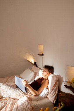 Kadın, yumuşak çarşaflar ve kırsal iç ayrıntılarla çevrili bir tabletle yatakta dinleniyor. Ilık sabah ışığı geleneksel trullo yatak odasına giriyor, güne huzurlu bir başlangıç yapıyor.