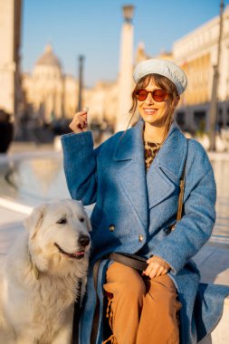 Mavi ceketli gülümseyen bir kadın St. Peter Bazilikası yakınlarındaki büyük beyaz köpeği yanında oturan modern bir çeşmeye bozuk para atıyor. Roma 'da umudun sembolü ve neşeli seyahat anları