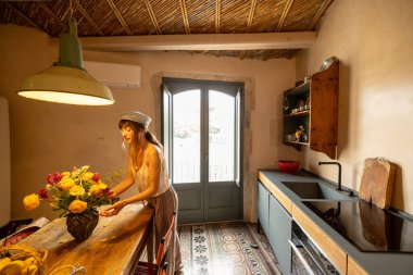 Geleneksel Sicilya tasarımı, desenli fayanslar ve açık kapılar ardında tarihi yamaç kenti manzarası olan güneşli kırsal bir mutfakta taze çiçekler düzenleyen bir kadın.