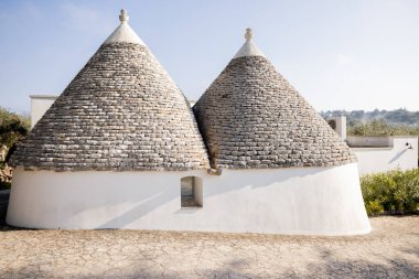 Güney İtalya 'da berrak bir gökyüzünün altında beyazlatılmış trullo evinin iki geleneksel konik çatısı sabah ışığında ikonik Apulian mimarisi sergiliyor.