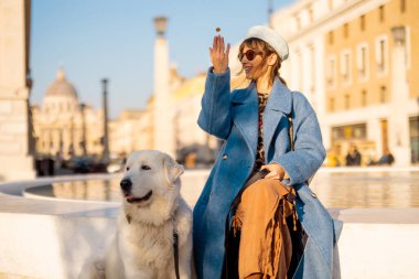 Mavi ceketli gülümseyen bir kadın St. Peter Bazilikası yakınlarındaki büyük beyaz köpeği yanında oturan modern bir çeşmeye bozuk para atıyor. Roma 'da umudun sembolü ve neşeli seyahat anları