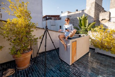 Neşeli bir kadın kendini güneşin altındaki bir çatıda kaydediyor. Etkilenen yaşam tarzı, dijital göçebeler, uzaktan çalışma veya sosyal medya pazarlaması gibi konular için harika