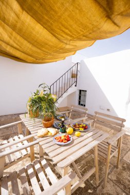 Beyaz bir terasta Rustik ahşap masa. Taze Akdeniz yemekleri, limonlar, çiçekler ve zeytinyağı ile altın sarısı bir güneş gölgesinin altında. Sıcak ve rahat yaz havası
