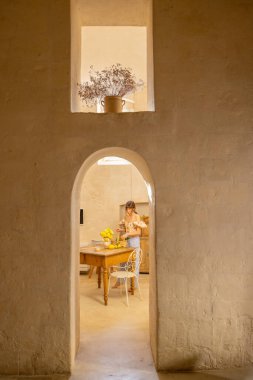 Bir kadın taştan bir mutfaktaki ahşap bir masada sarı çiçekler düzenliyor. Sahne basitlik, gelenek ve konforlu Akdeniz yaşamını iç mekan veya yaşam tarzı kullanımı için mükemmel bir şekilde yansıtıyor