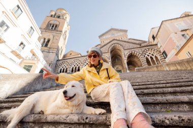 Bir kadın ve köpeği sabah güneşinin altında Amalfi 'deki katedralin merdivenlerinde dinleniyorlar. Bu fotoğraf, ikonik bir İtalyan ortamında boş zamanları, arkadaşlığı ve otantik seyahat anlarını yakalar.