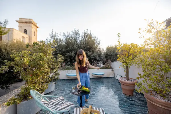 Güneşli terasta canlı bir buket sebze ve çiçek tutan bir kadın. Beyaz bir köpek, çizgili tekstil, narenciye ve kırsal taş mimarisi ile rahat bir ortam.