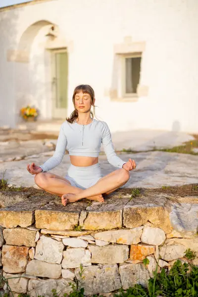 Güney İtalya 'da geleneksel trullo' nun önünde yoga yapan bir kadın. Dengeleyici bir duruşta, doğayla ve sabah ışığıyla bağlantı kurar, kendini antik taşlara yaslar.