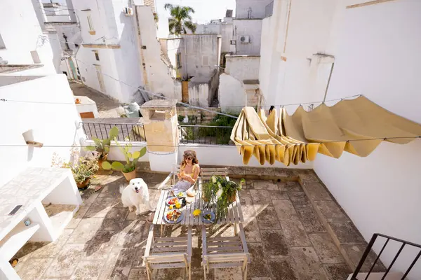 Güney İtalya 'da kırsal çatı terasında taze meyve ve kahveyle yavaş yavaş kahvaltı yapan bir kadın. Yaz ışığı, kaktüs bitkileri ve tüylü köpek dingin sabah havasını tamamlıyor.