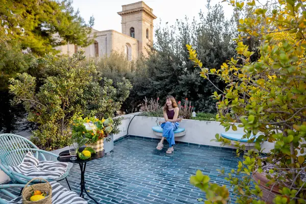 Bir kadın, bahçenin çatısındaki terasta, bitki ve tarihi Akdeniz mimarisi ile çevrili, huzurlu ve yavaş bir yaşam tarzını çağrıştıran, sessiz bir anın tadını çıkarır.