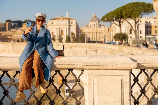 Açık mavi ceketli bir kadın Roma köprüsünde neşeli bir selfie çekiyor ve arka planda St. Peters Bazilikası sabah güneşinin altında parlıyor.