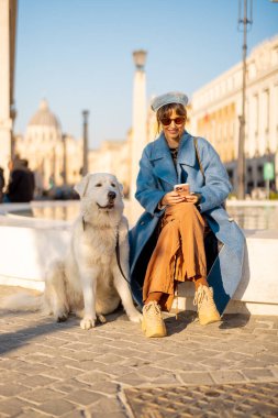 Vatikan 'ın yanındaki çeşme kenarında oturan bir kadın büyük beyaz köpeğiyle telefonuyla gülümsüyor. Modern seyahat yaşam tarzı ve evcil hayvan dostu turizm temaları için mükemmel bir imaj.