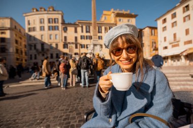 Mavi ceketli bir kadın sabah kahvesini Roma 'daki Pantheon' un önündeki masada içiyor. Güneşin ve hayat dolu sokak hayatının tadını çıkarıyor.
