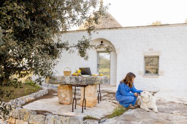 Mavi tulumlu bir kadın, köpeği ve laptopuyla geleneksel bir trullo 'nun önünde güvenle duruyor. Konsept iş ve kırsal yaşam arasındaki dengeyi gösteriyor.