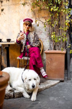 Klasik kıyafetli bir kadın büyük beyaz köpeğiyle Roma 'da büyüleyici bir kafede oturuyor. Görüntü, rahat şehir yaşam tarzını seyahat için mükemmel yansıtıyor.