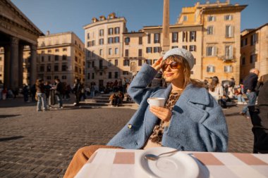 Mavi ceketli bir kadın sabah kahvesini Roma 'daki Pantheon' un önündeki masada içiyor. Güneşin ve hayat dolu sokak hayatının tadını çıkarıyor.