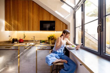 Bir kadın, güneşli bir mutfakta sağlıklı bir kahvaltı yaparken video görüşmesinde el sallıyor. Uzak iş, yaşam tarzı teknolojisi ya da modern yaşam hakkında reklamlar için ideal.