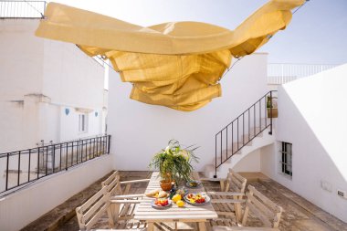 Beyaz bir terasta Rustik ahşap masa. Taze Akdeniz yemekleri, limonlar, çiçekler ve zeytinyağı ile altın sarısı bir güneş gölgesinin altında. Sıcak ve rahat yaz havası