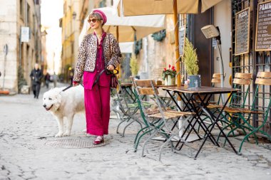 Kalın renkli şık bir kadın tüylü beyaz köpeğiyle büyüleyici bir Roma kafesinin önünden geçiyor. Sahne, sakin bir şehir gezisi ve İtalya 'daki günlük hayatın zarafetini yakalıyor.