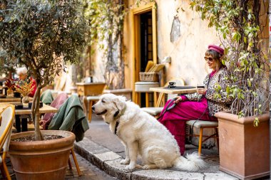 Klasik kıyafetli bir kadın büyük beyaz köpeğiyle Roma 'da büyüleyici bir kafede oturuyor. Görüntü, rahat şehir yaşam tarzını seyahat için mükemmel yansıtıyor.