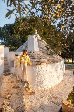Puglia 'da zeytin ağaçları ve yeşilliklerle çevrili güneşli bir trullo' nun yanında yürüyen bir kadın. Bu resim seyahat refahı veya kırsal yaşam kavramlarında kullanmak için miras ve yaşam tarzını harmanlıyor