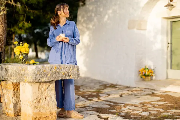 Yumuşak mavi keten giymiş bir kadın, güney İtalyan trullo 'sunun sakin güzelliğiyle çevrili sarı çiçeklerle bezenmiş taştan bir masanın yanında sakin bir sabah kahvesinden hoşlanır.