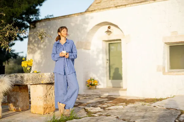 Yumuşak mavi keten giymiş bir kadın, güney İtalyan trullo 'sunun sakin güzelliğiyle çevrili sarı çiçeklerle bezenmiş taştan bir masanın yanında sakin bir sabah kahvesinden hoşlanır.