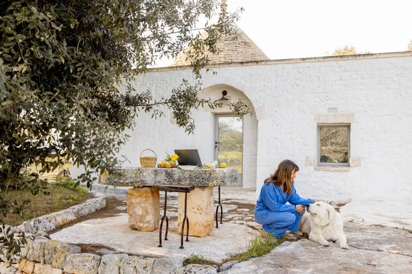 Mavi tulumlu bir kadın, köpeği ve laptopuyla geleneksel bir trullo 'nun önünde güvenle duruyor. Konsept iş ve kırsal yaşam arasındaki dengeyi gösteriyor.