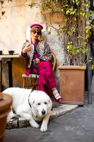 Klasik kıyafetli bir kadın büyük beyaz köpeğiyle Roma 'da büyüleyici bir kafede oturuyor. Görüntü, rahat şehir yaşam tarzını seyahat için mükemmel yansıtıyor.