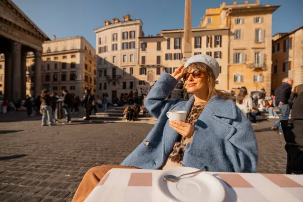Mavi ceketli bir kadın sabah kahvesini Roma 'daki Pantheon' un önündeki masada içiyor. Güneşin ve hayat dolu sokak hayatının tadını çıkarıyor.