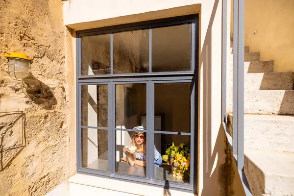 Güneş gözlüklü ve keten elbiseli bir kadın elinde telefonla güneşli bir pencere pervazında oturuyor. Yaşam tarzı blogları, moda yazıları ya da yavaş yaşam kampanyaları için mükemmel.