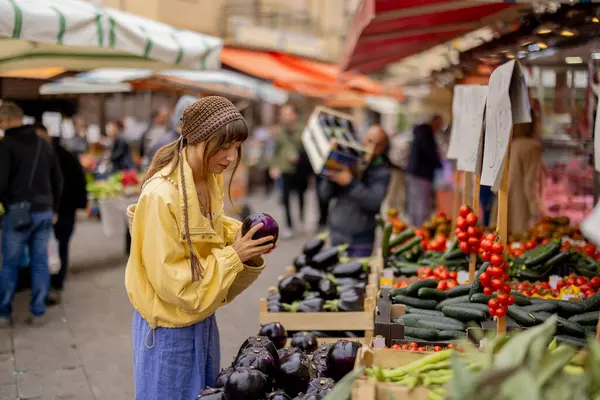 Sarı ceketli bir kadın canlı bir sokak pazarından patlıcan seçiyor. Bu görüntü dikkatli bir yaşamı, sağlıklı seçimleri ve yerel yemek kültürünün cazibesini yansıtıyor.