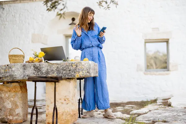 Mavi elbiseli bir kadın dışarıda, taşra bir masada dizüstü bilgisayarın yanında dikilirken görüntülü görüşme sırasında el sallıyor. Resim, yavaş yaşam ve bağlantı dokunuşuyla uzaktan çalışmayı gösteriyor