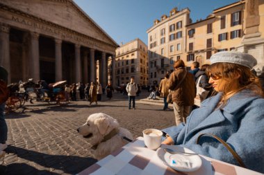 Mavi ceketli bir kadın sabah kahvesini Roma 'daki Pantheon' un önündeki masada içiyor. Güneşin ve hayat dolu sokak hayatının tadını çıkarıyor.