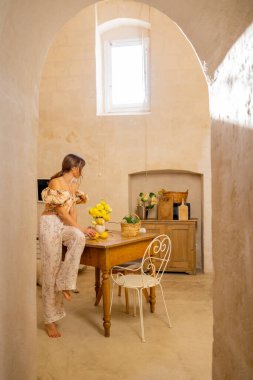 Bir kadın taştan bir mutfaktaki ahşap bir masada sarı çiçekler düzenliyor. Sahne basitlik, gelenek ve konforlu Akdeniz yaşamını iç mekan veya yaşam tarzı kullanımı için mükemmel bir şekilde yansıtıyor