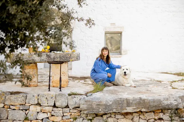Mavi elbiseli bir kadın İtalya 'da geleneksel trullo' nun önünde beyaz köpeğini nazikçe okşar. Fotoğraf, güven, huzurlu kırsal yaşam tarzı ve hayvanlarla duygusal bağlantıyı ifade ediyor.