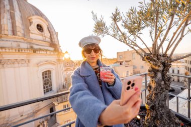 Roma 'nın çatısında elinde kırmızı bir kokteyl tutarak selfie çeken bir kadın. Kubbe ve zeytin ağacı bu canlı şehre cazibe katıyor.