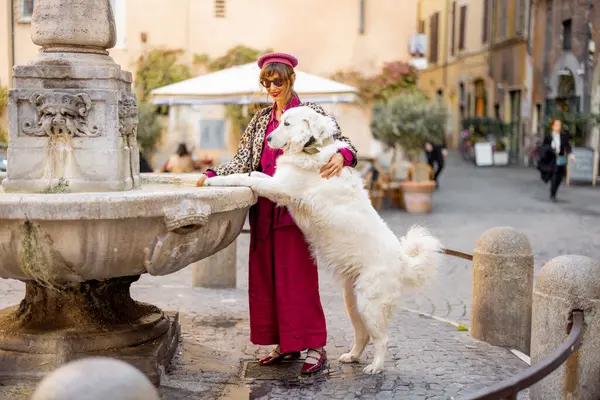 Roma 'da tarihi bir fıskiyenin yanında şık kıyafetler içinde gülümseyen bir kadın ve büyük beyaz bir köpek duruyor. Bu fotoğraf özgün bir Avrupa cazibesi ve güçlü bir insan hayvan bağı taşır.
