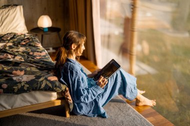 Mavi keten giysili bir kadın panoramik pencerenin yanında oturup tablet okuyor. Yumuşak sabah ışığı, rahat bir atmosfer ve doğayla barışçıl bir bağ.