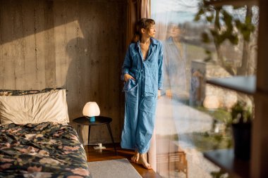 Bir kadın, rahat bir yatak odasında, büyük bir pencerenin yanında çıplak ayakla sabah güneşinin tadını çıkarıyor. Bir durgunluk, sıcaklık ve doğal güzellik anı.