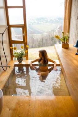 Bir kadın panoramik pencerenin yanında, tahta bir küvette huzurlu bir banyo yapmaktan hoşlanır. Çiçeklerle ve gün ışığıyla çevrili bu an, huzurlu bir kişisel bakım ve sessiz bir lüksü yakalar.