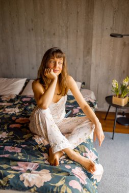 Çiçekli pijamalı bir kadın yatakta oturup düşüncelere dalmış. Yavaş yaşam, farkındalık, sabah ritüelleri veya başyazı ve ticari içeriklerde rahat ev modasını teşvik etmek için ideal