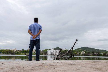 Genç adam, sabah dramatik gökyüzü ile el değmemiş sakin gölü seyrediyor.