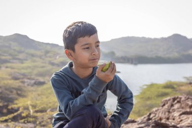 İzole edilmiş genç bir çocuk, sabahları göl manzaralı bir dağ tepesinde guava yiyor.