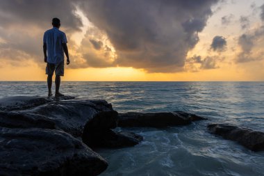 Kala Pathar Sahili 'nde Andaman ve Nicobar Adaları' nı kasıp kavuran kayalık kıyılarda ve şafak vakti dramatik gökyüzü ile deniz ufkuna bakan yalıtılmış genç adam görülüyor..