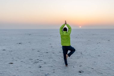 young man performing yoga in white desert with sunrise orange dramatic sky at dawn image is taken at dhordo kutch white rann gujrat india. clipart