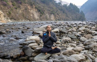 Genç adam sabah sakin bir dağ nehri yatağında taşın üzerinde meditasyon yapıyor.