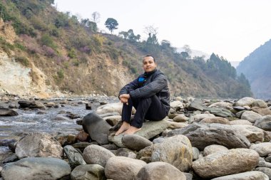 Bir adam sakin bir dağda nehir yatağında oturmuş sabah doğayı seyrediyor.