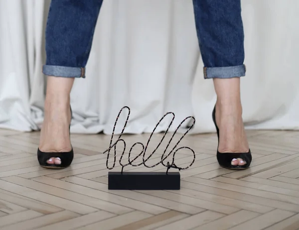 Close-up of female legs in blue jeans and black high-heeled shoes against a background of beige drapery, the inscription Hello between the legs. Womens leather shoes.selective focus