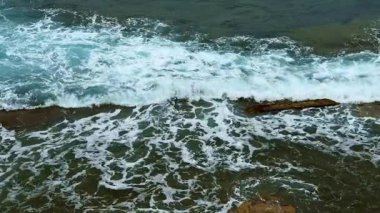 Okyanus dalgaları engebeli kayalara çarparak beyaz köpük ve çağlayan su oluşturur. Deniz güçlü bir şekilde kıyı şeridini yutar, dinamik ve büyüleyici bir doğal manzara oluşturur. Üst görünüm